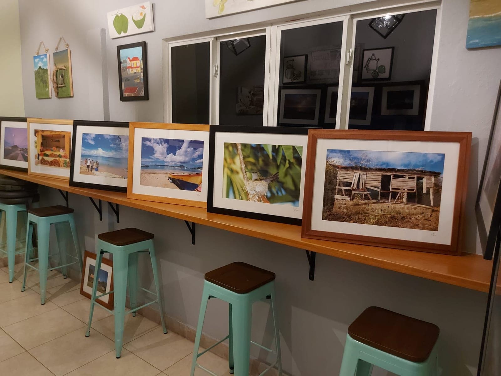 A row of framed photographs propped along the café’s counter shelf for the art show — beach, boats, a hummingbird at its nest and an old chattel house — with paintings hung on the wall behind