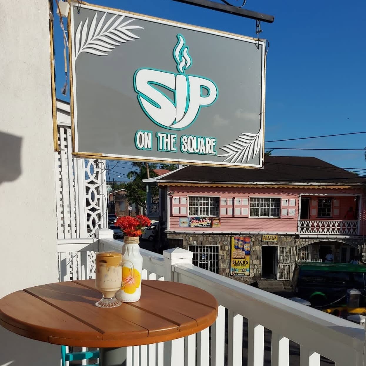 A balcony table set with an iced coffee, the café sign hanging overhead against a clear sky
