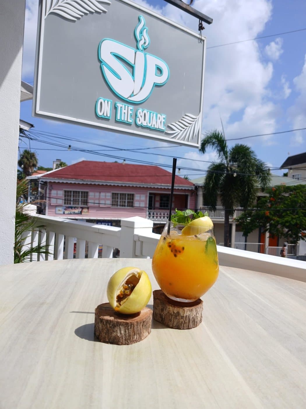 A passion fruit cooler with mint and a slice of lime on the veranda table, the SIP ON THE SQUARE sign hanging above and Charlestown behind