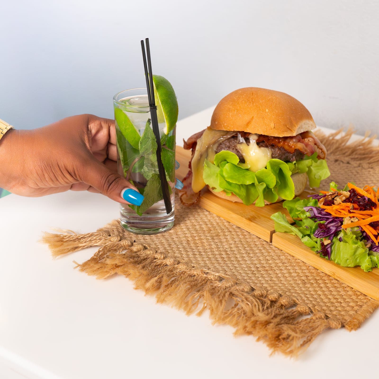 The Cowboy burger and slaw on a wooden board, a mojito set beside it on a burlap runner
