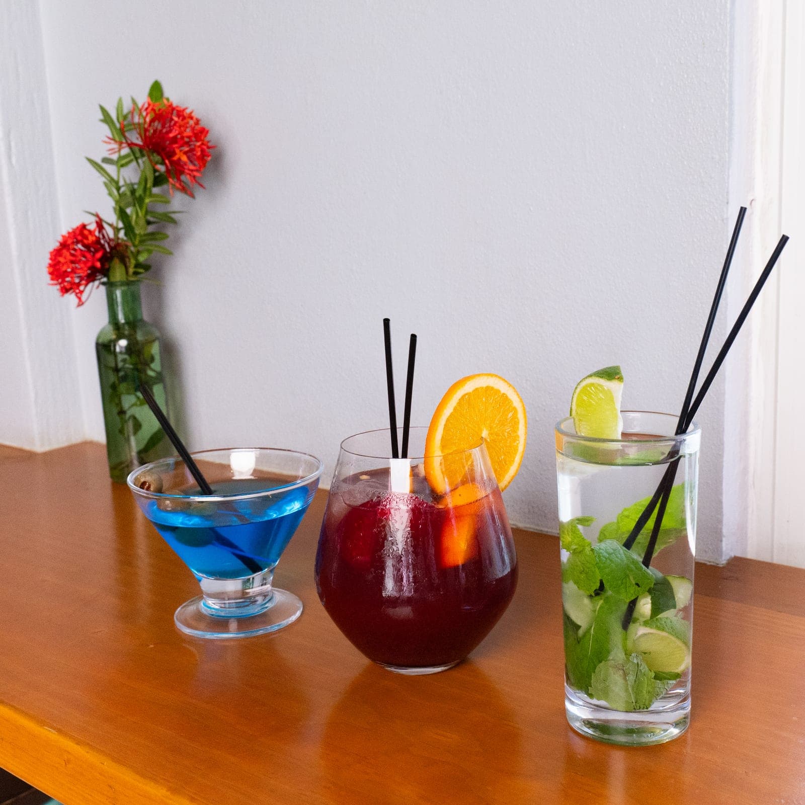 A blue cocktail, a fruit punch and a mojito lined up on a table, ixora flowers behind