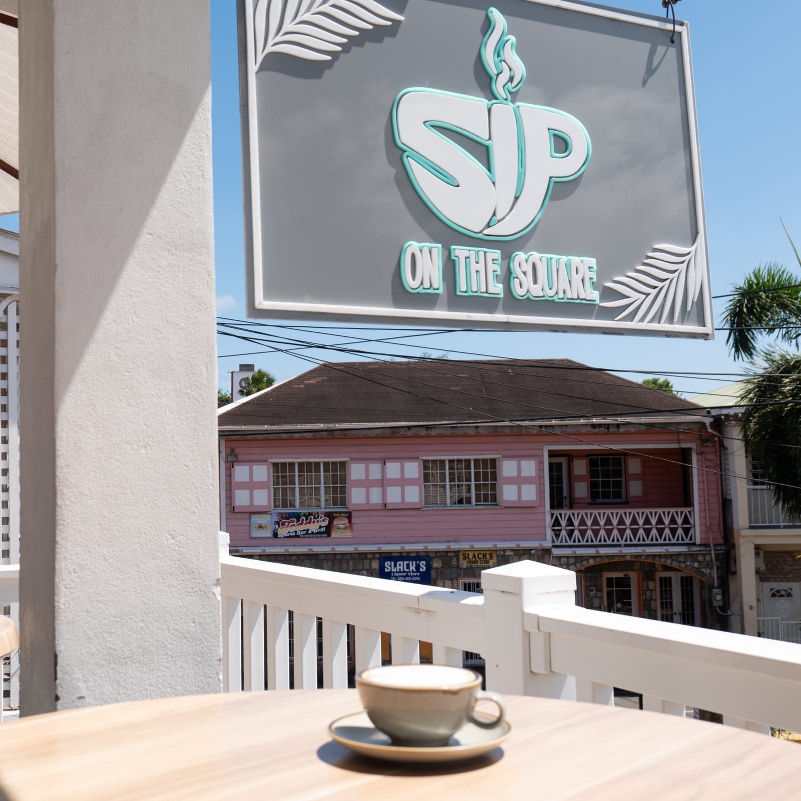A cappuccino on the veranda rail beneath the SIP ON THE SQUARE sign, Charlestown behind