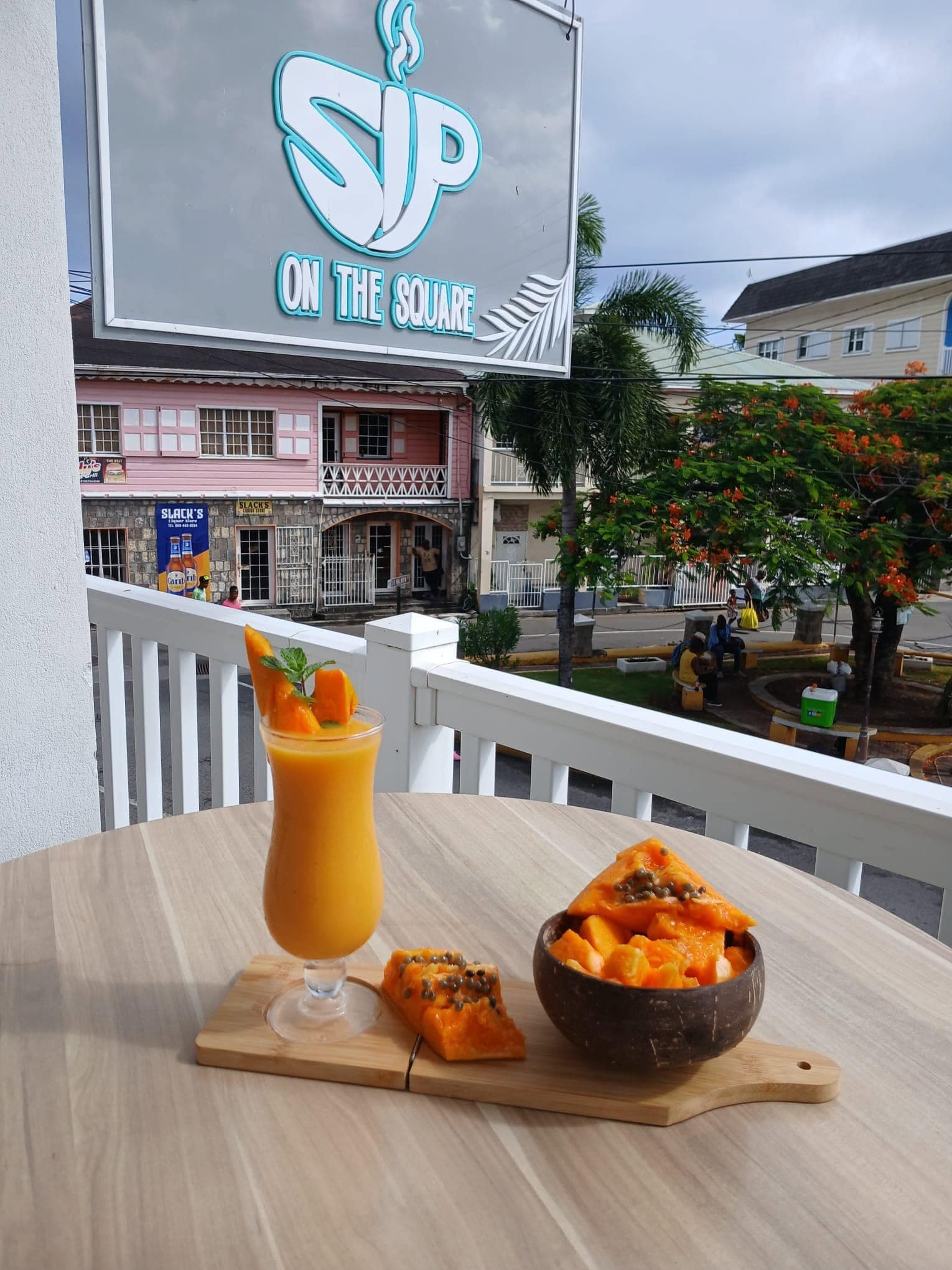 The SIP ON THE SQUARE sign above a veranda table set with a fresh fruit smoothie and a wooden board of sliced tropical fruit, looking out over Charlestown’s square